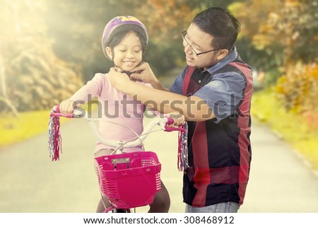 Young father helps his daughter to fasten helmet on the head while riding a bike on the park