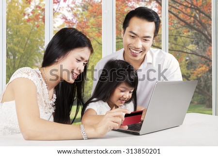 Cheerful parents and their daughter using laptop and credit card for shopping online at home with autumn background on the window