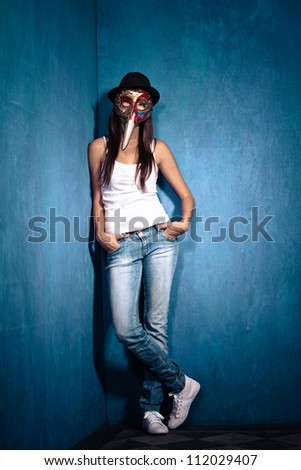 teen girl wearing Venetian mask,black hat and blue jeans stand in corner of room full body shot