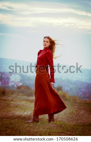 young blonde woman in long red skirt and leather jacket stand in the wind on hill above the city