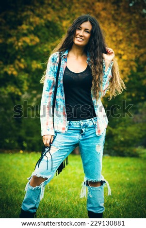 smiling young woman with long curly hair in blue torn jeans and tartan shirt outdoor shot autumn day in park
