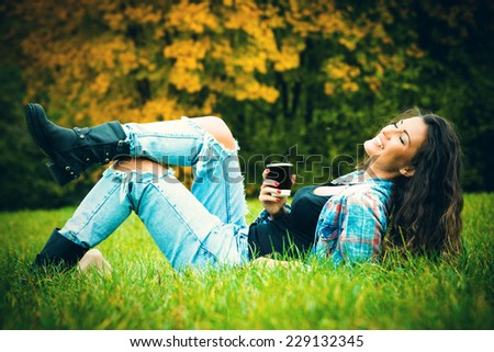 smiling young woman enjoy in coffee break in nature, lie on grass in park, wearing blue torn jeans and tartan shirt, full body shot