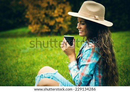 smiling young woman enjoy in coffee break in nature, sit in grass in park, wearing hat, blue torn jeans and tartan shirt