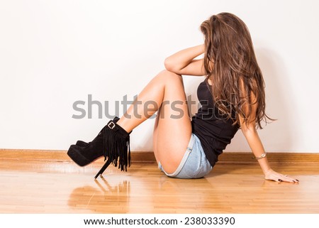 fashion girl in high heel shoes jeans shorts and black top tank sit on parquet against wall in room