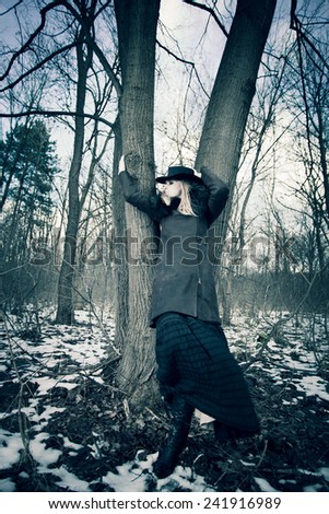 blonde woman wearing hat, leather coat and long skirt stand by tree  in the forest, cold winter day, full body shot