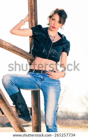 young rebel woman in blue jeans, leather boots and leather jacket outdoor shot on old metal construction hot sunny day