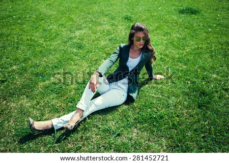elegant young woman with sunglasses, green jacket,  white pants and high heel shoes sit on grass in park, full body shot