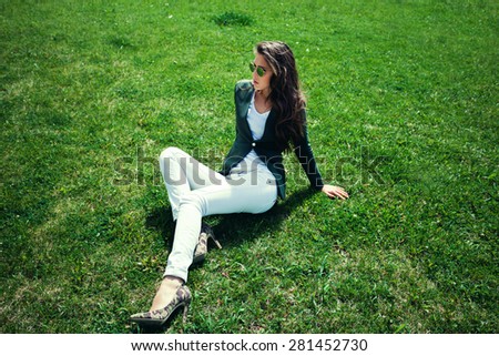 elegant young woman with sunglasses, green jacket,  white pants and high heel shoes sit on grass in park, full body shot
