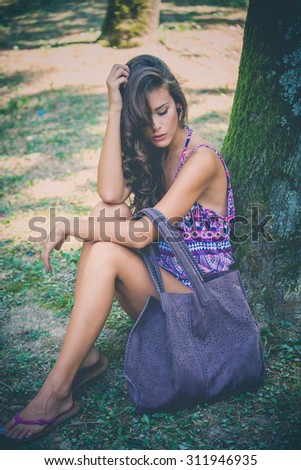 young beautiful woman sit by tree in park wearing purple print top and big leather bag, full body shot