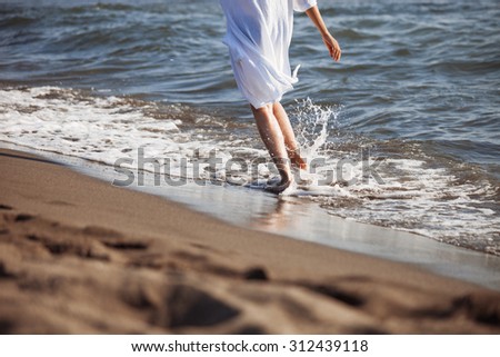 barefoot woman in white shirt run through sea water on sandy beach, back view, lower body