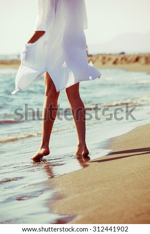barefoot woman enjoy in sea water on sandy beach in white long shirt, lower body, back view