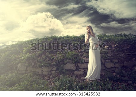 young blonde woman in long white dress with twig of flowers in hand stand by old wall overgrown with grass, outdoor shot, full body shot