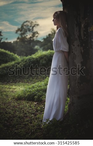 young woman in long white dress lean against big tree looking far away, full body shot, profile, summer day