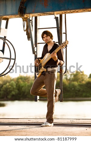 young man play bass guitar at industrial area by the river at sunset, full body shot