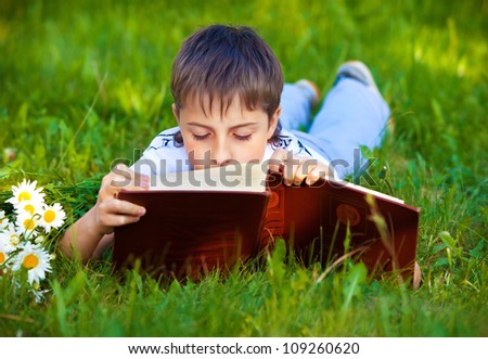 young pupil boy reading a book while lying on green grass in the park