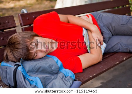 young man traveler resting on bench at park. He holds map on hands. There is backpack under his head
