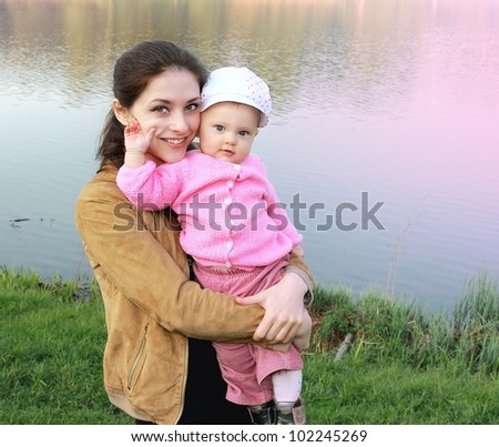 Beautiful mather holding baby girl with love in pink dress and funny hat on nature lake and green grass background. Baby holding mother cheek
