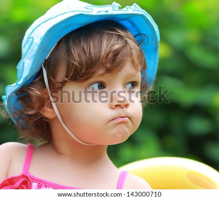 Angry baby girl looking in blue hat on summer background. Closeup portrait