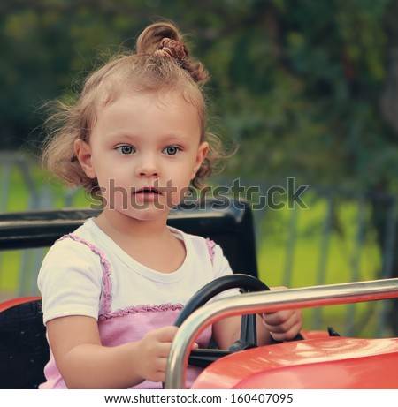 Funny thinking kid girl driving car in recreation park on summer background. Closeup portrait