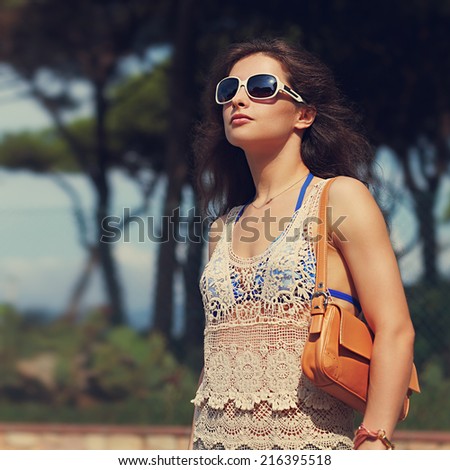 Beautiful urban woman in beach dress and sunglasses with leather bag on shoulder outdoors summer background. Closeup vintage portrait