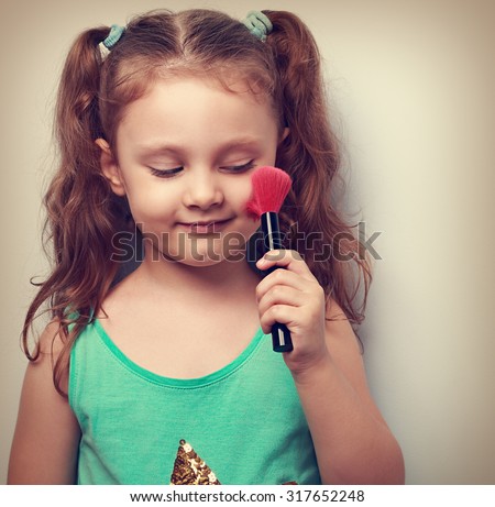 Fun beautiful small kid girl applying tonal cosmetic on the face using makeup brush with happy smile. Closeup portrait