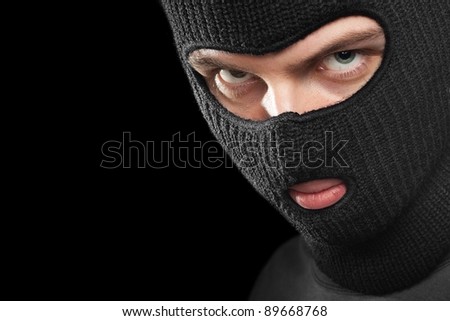 Evil criminal in a mask looking at the viewer; isolated on black background, copy space on the left.