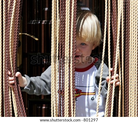 Child or young boy with blond or blonde hair and blue eyes looking out from behind curtain with a cheeky look on his face