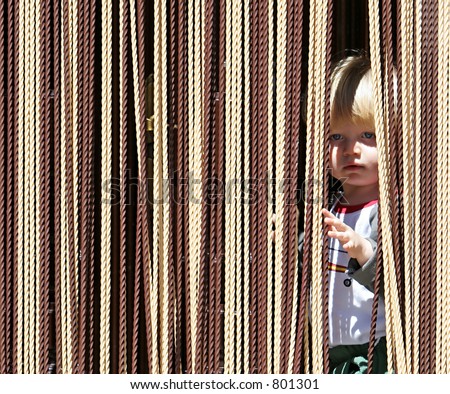Child or young boy with blond or blonde hair and blue eyes looking out from behind curtain with a cheeky look on his face.