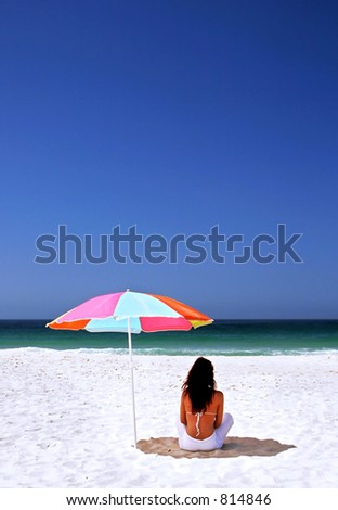 Young, attractive, fit, tanned woman sitting in shade on Spanish beach under sun umbrella. White sand blue sea and sky.