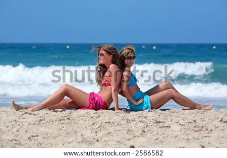 Two young attractive bikini clad women chilling on a sunny beach on holiday or vacation by the sea