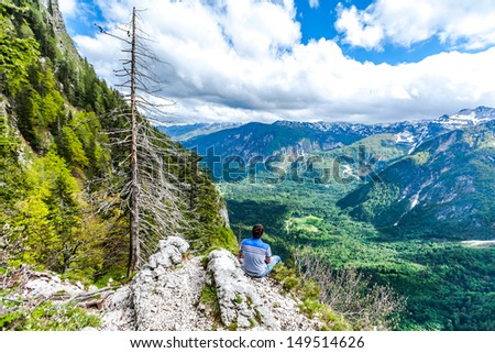 The man sitting on the cliff`s edge on the wide mountains landscape background. The Julian Alps, Slovenia. View on the Bohinj Lake.