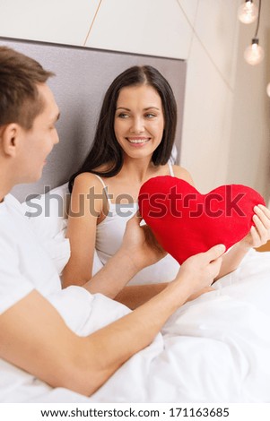 hotel, travel, relationships, holidays and happiness concept - smiling couple in bed with red heart-shaped pillow