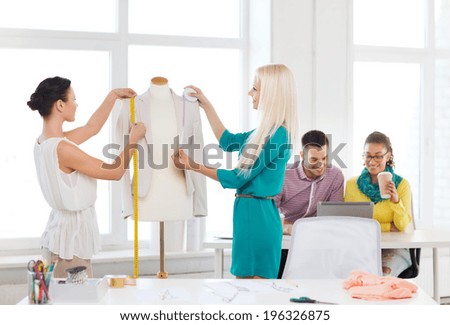 startup, education, fashion and office concept - smiling fashion designers measuring and decorating jacket on mannequin