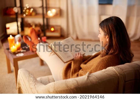 halloween, holidays and leisure concept - young woman reading book and resting her feet on table at cozy home