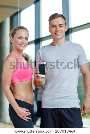 fitness, sport, advertising, technology and diet concept - smiling young woman and personal trainer with smartphone blank screen in gym