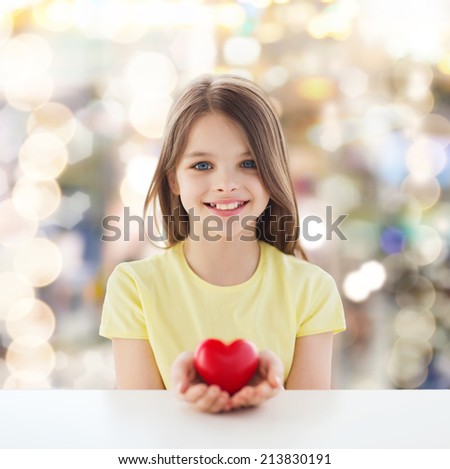 childhood, love, charity, holidays and people concept - smiling little girl sitting and holding red heart over sparkling background