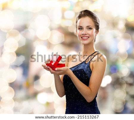 holidays, presents, luxury and happiness concept - smiling woman in dress holding red gift box over lights background