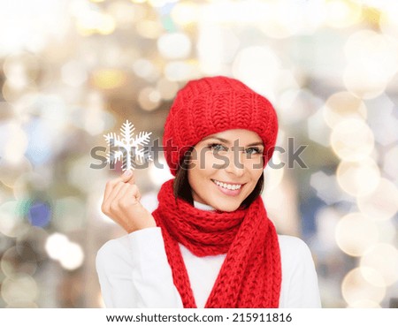 happiness, winter holidays, christmas and people concept - smiling young woman in red hat, scarf and mittens holding snowflake over lights background