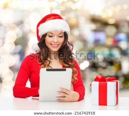 christmas, holidays, technology and people concept - smiling woman in santa helper hat with gift box and tablet pc computer over lights background