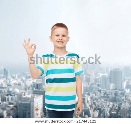 childhood, gesture and people concept - smiling little boy in casual clothes making ok gesture over city background