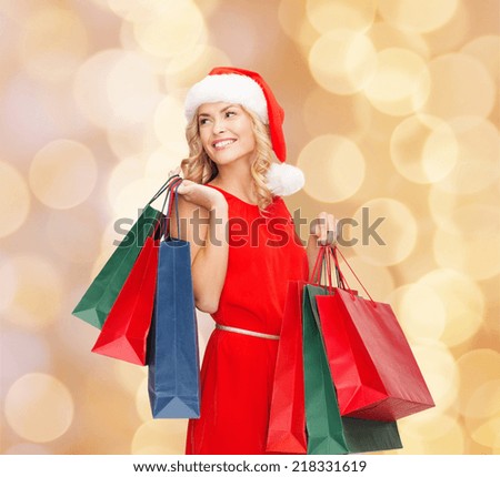 sale, gifts, christmas, holidays and people concept - smiling woman in red dress and santa helper hat with shopping bags over beige lights background