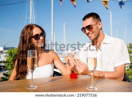 love, dating, people and holidays concept - smiling couple wearing sunglasses with champagne and small red gift box putting wedding ring on finger at cafe
