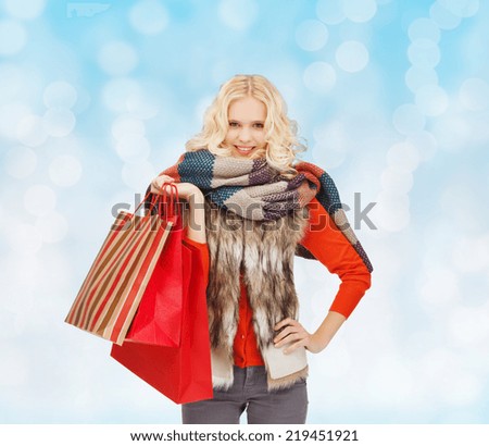 happiness, winter holidays, christmas and people concept - smiling young woman in winter clothes with red shopping bags over blue lights background