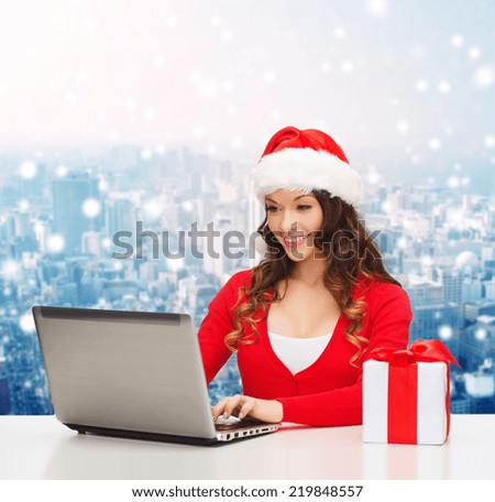 christmas, holidays, technology and people concept - smiling woman in santa helper hat with gift box and laptop computer over snowy city background