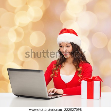 christmas, holidays, technology and people concept - smiling woman in santa helper hat with gift box and laptop computer over biege lights background