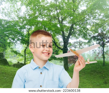 dreams, future, hobby, nature and childhood concept - smiling little boy holding wooden airplane model in his hand over park background