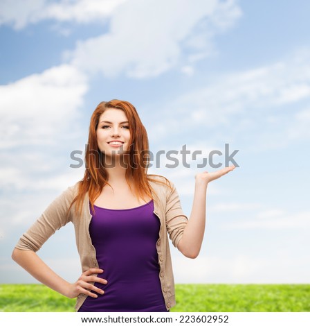 nature, summer, advertising and people concept - smiling teenage girl in casual clothes holding something on her palm over blue sky and grass background
