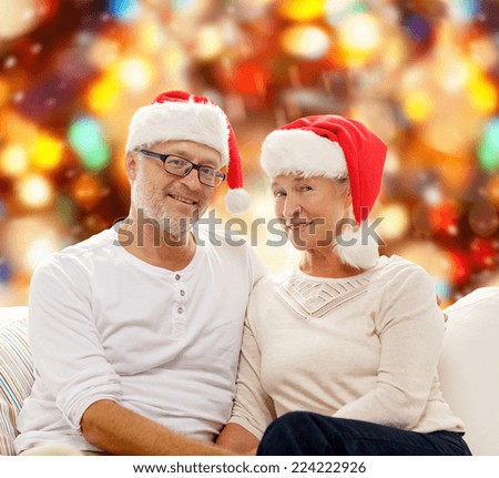 family, holidays, christmas, age and people concept - happy senior couple in santa helper hats sitting on sofa over red lights background