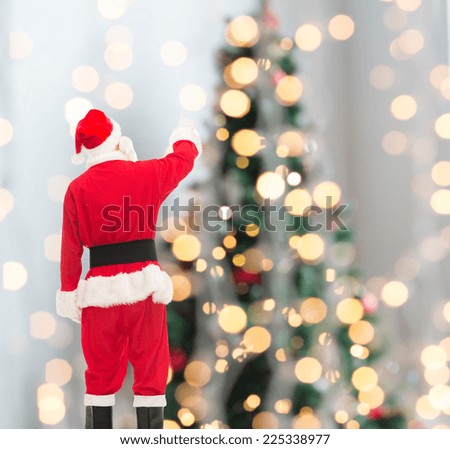 christmas, holidays and people concept - man in costume of santa claus pointing finger from back over tree lights background