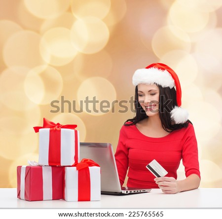 christmas, shopping and people concept - smiling woman in santa helper hat with gift boxes, laptop computer and credit card over beige lights background
